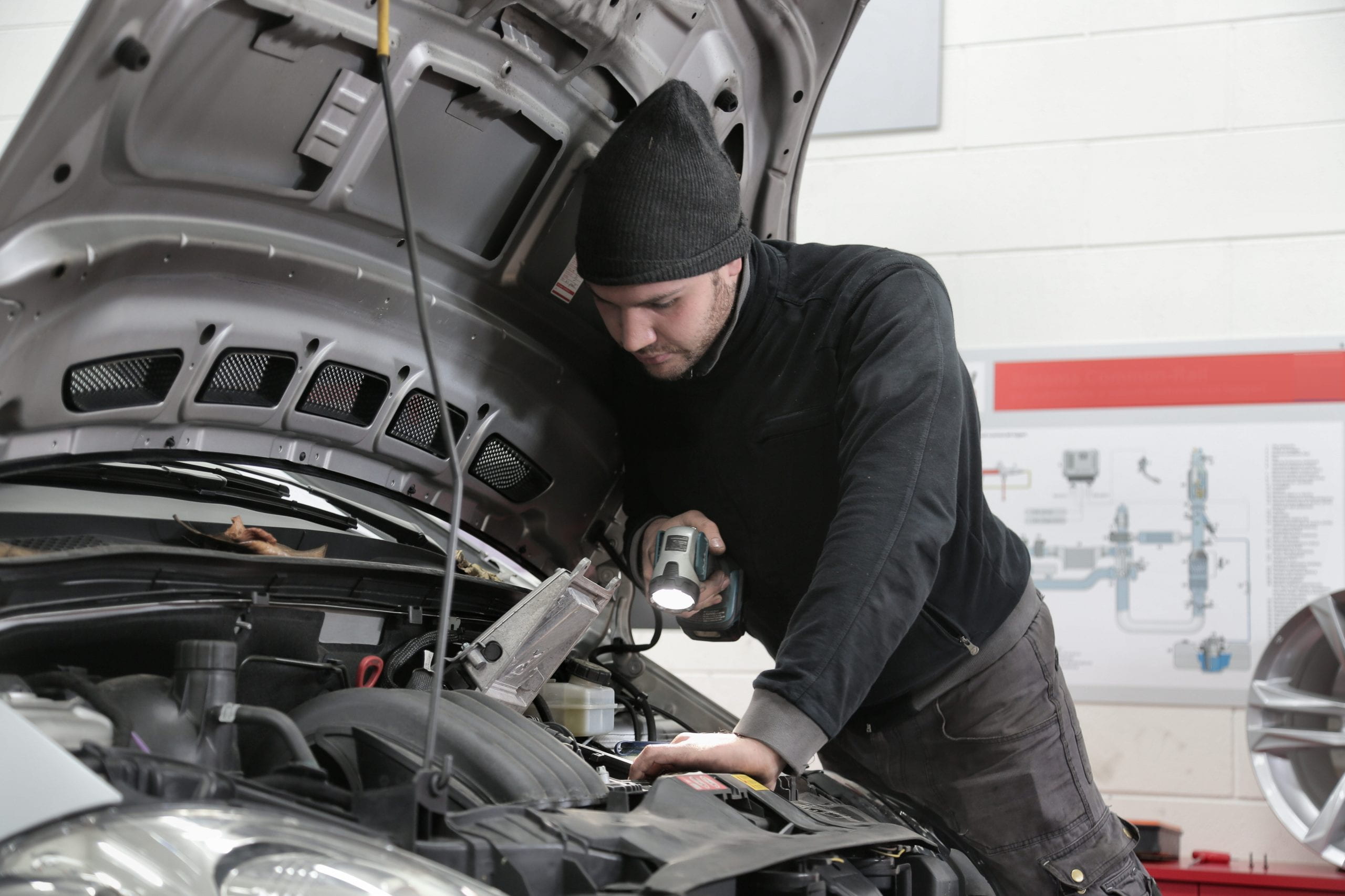 A young male in black clothes and a hat, conducting an MOT on a car with an open bonnet.